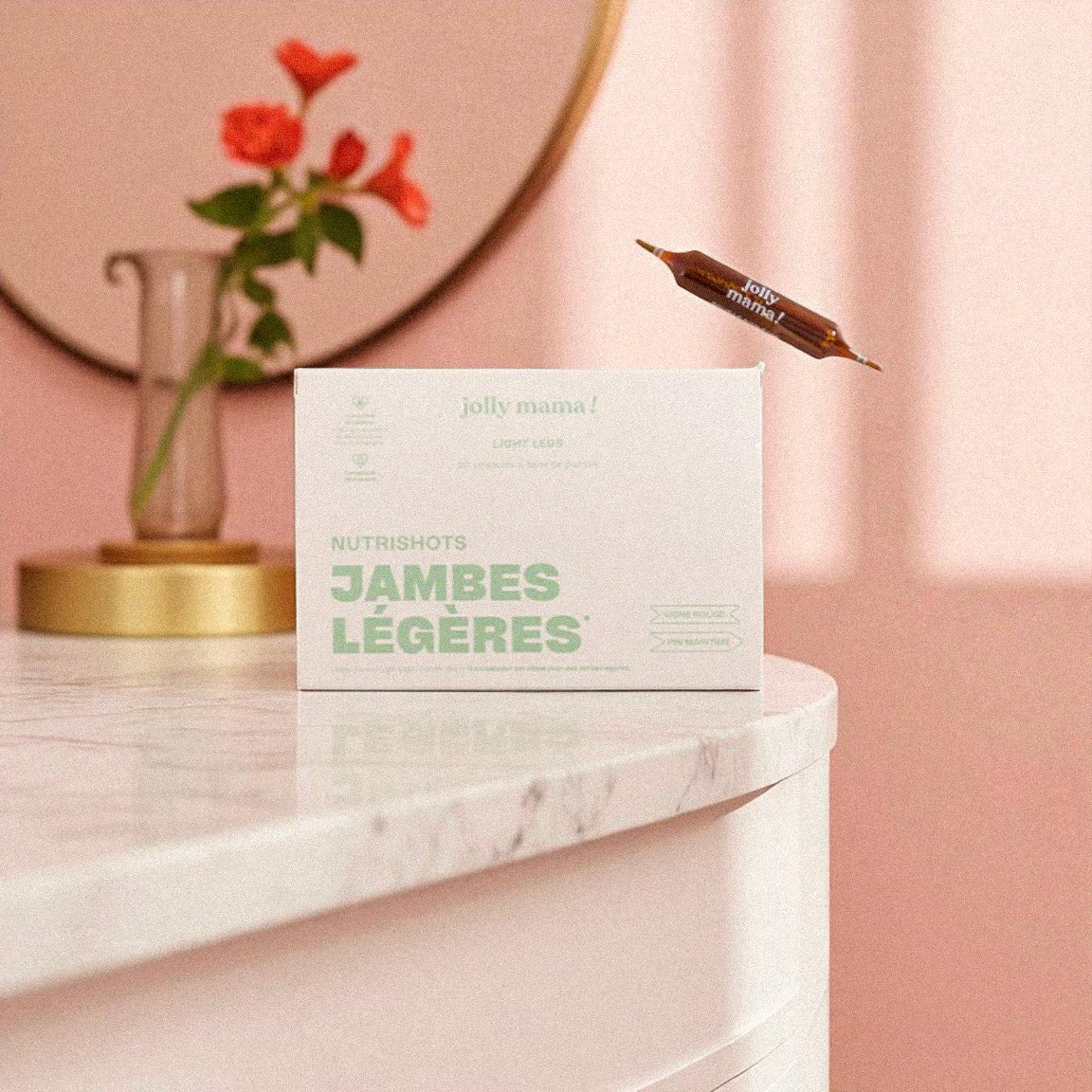 Packshot du complément alimentaire Jambes Légères Jolly Mama avec une ampoule posée sur une table en marbre et un vase fleuri.