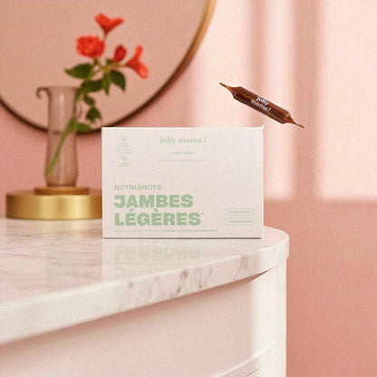 Packshot du complément alimentaire Jambes Légères Jolly Mama avec une ampoule posée sur une table en marbre et un vase fleuri.