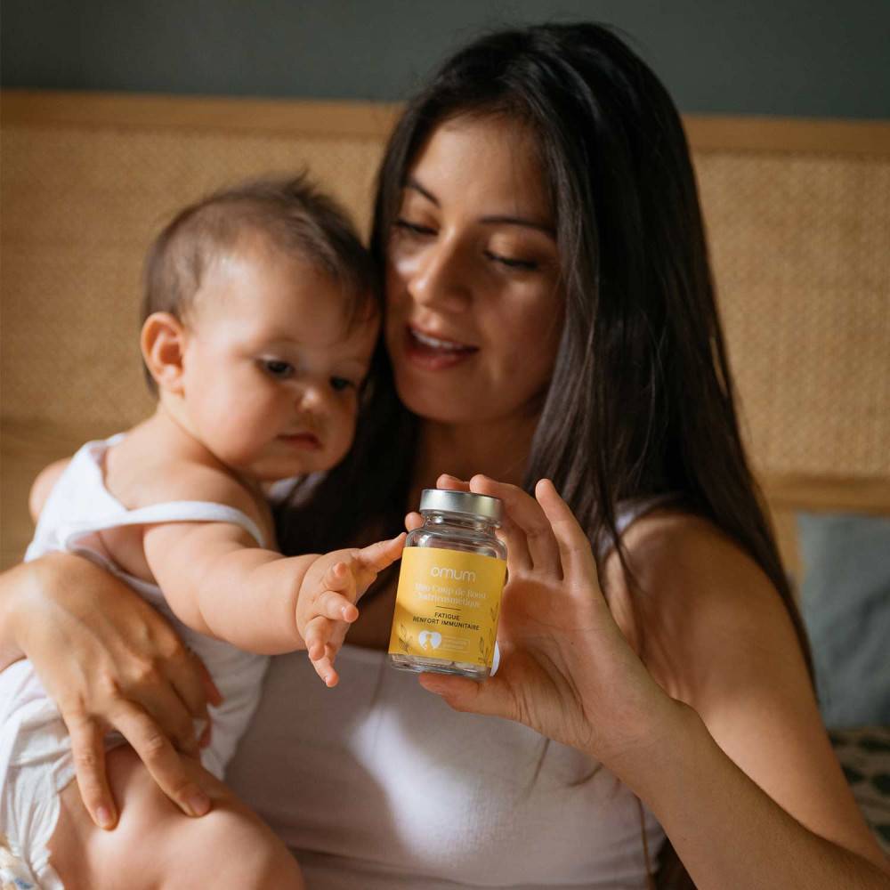 Jeune maman avec son bébé tenant le pilulier Omum Mon Coup de Boost, complément alimentaire naturel compatible grossesse et allaitement.